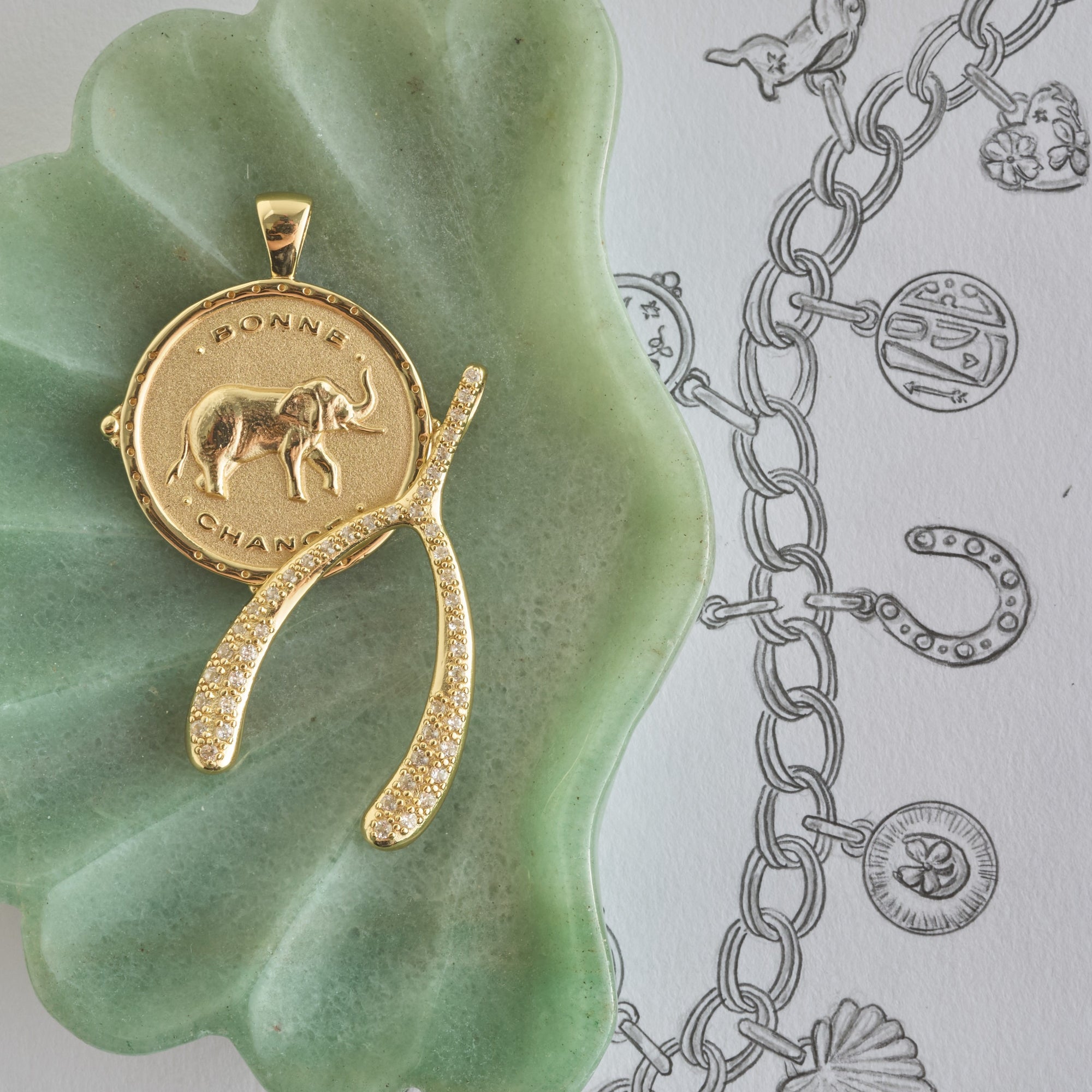 Stylized shot of bonne chance elephant coin and lucky pave wishbone in green dish on a sketch of jewelry
