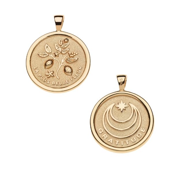 Front of gold coin pendant features a branch with lemons and lemon blossoms and the phrase "LA PLUS BELLE FLEUR" and back of pendant features moon and star logo with the word "GRATITUDE"