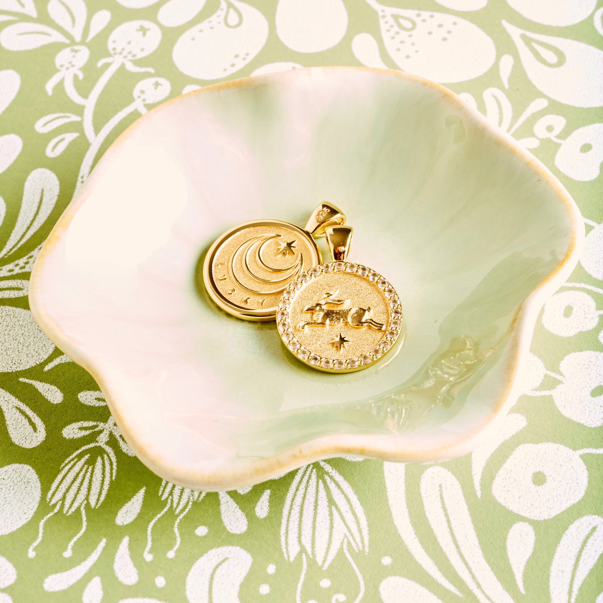 Stylized shot showing the front and back of the lucky petite embellished coin in a light green ceramic dish