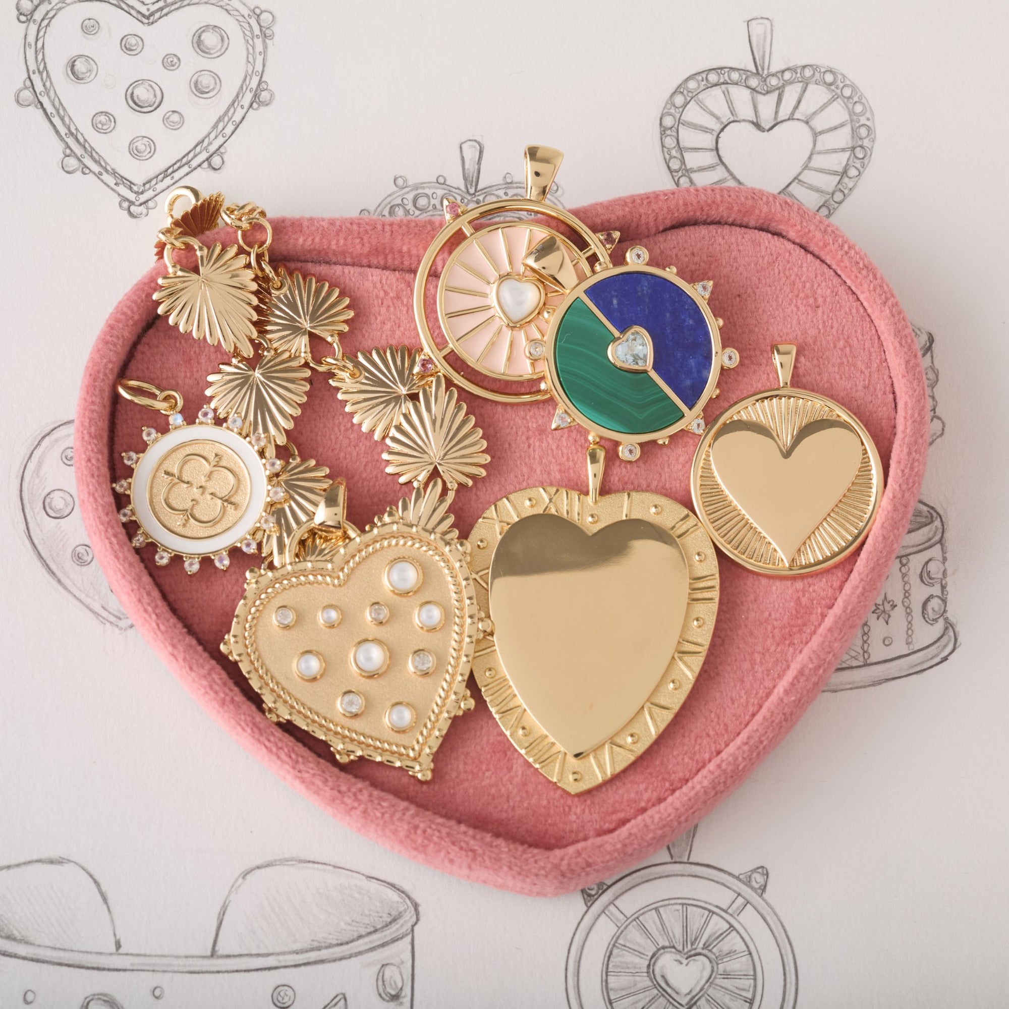 Heart shaped tray filled with assorted love-themed pendants and bracelets, sitting on top of heart sketches.