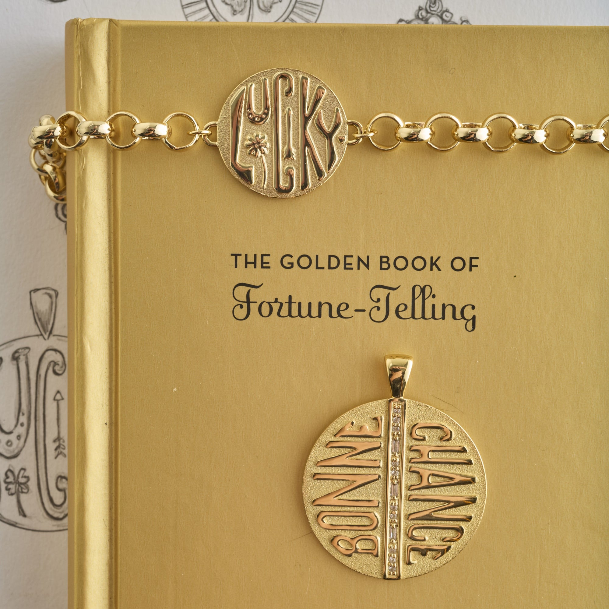 Stylized show of jumbo bonne chance coin and bonne chance coin bracelet on a gold book