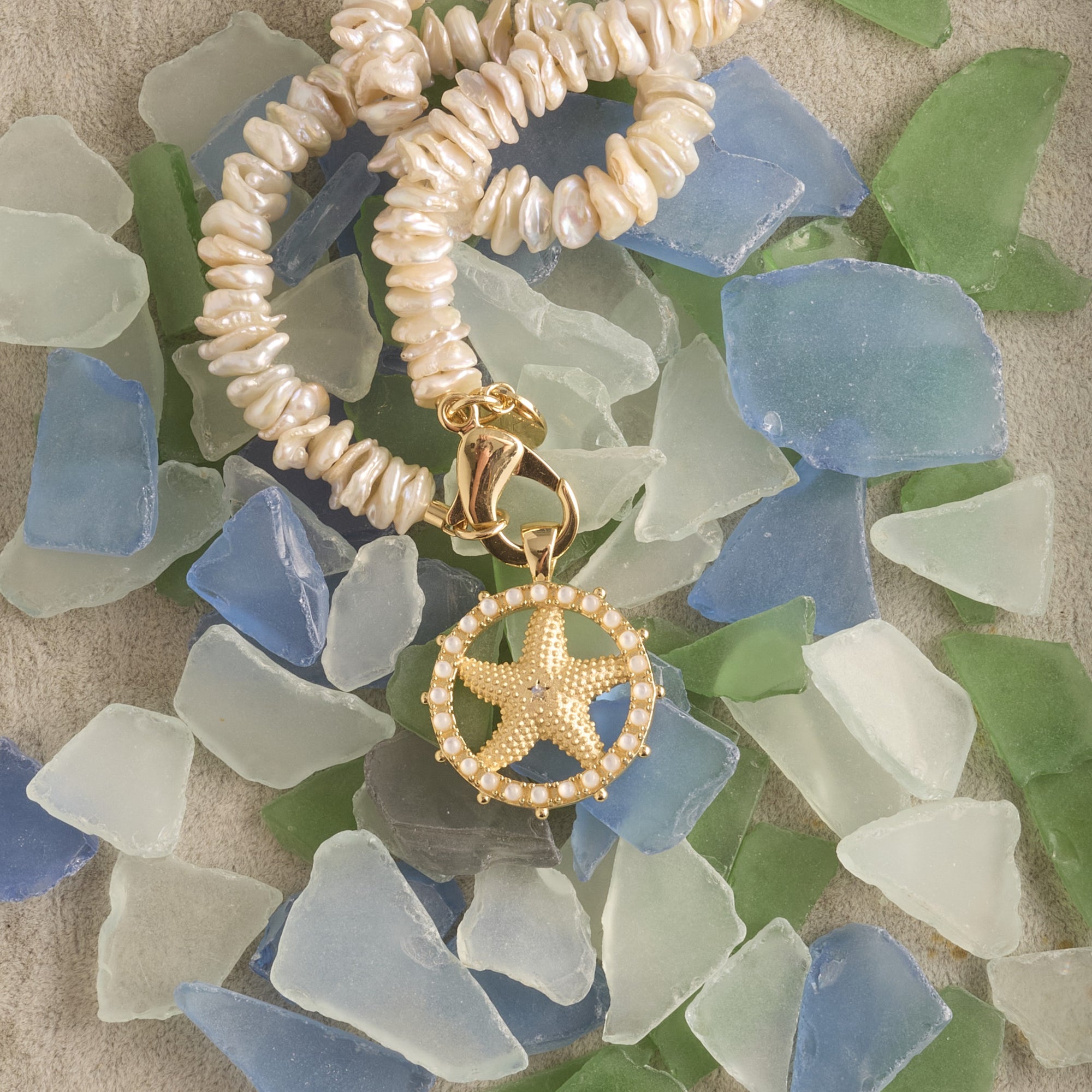 Stylized shot of starfish cutout coin on mother of pearl beaded necklace laying on a pile of sea glass 