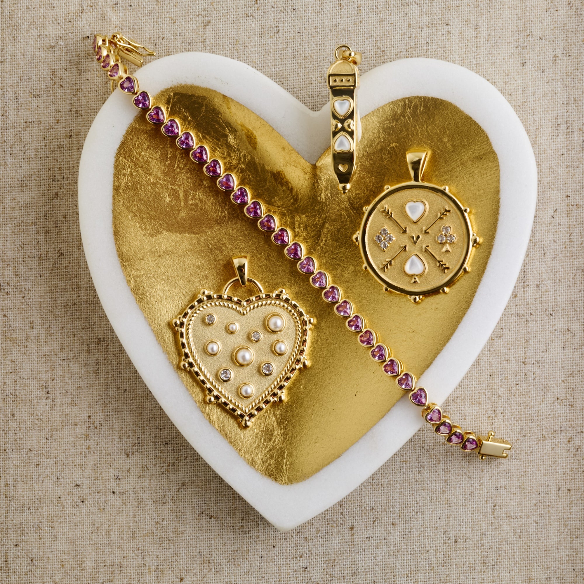 Stylized shot of the queen of hearts coin pendant, heart ray pendant, love in the air heart pendant, and made from love tennis bracelet in gold heart dish